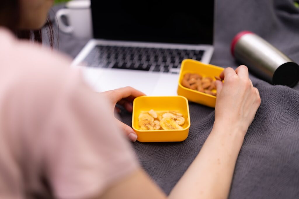 A person eating dried fruit and nuts from yellow bowls near a laptop